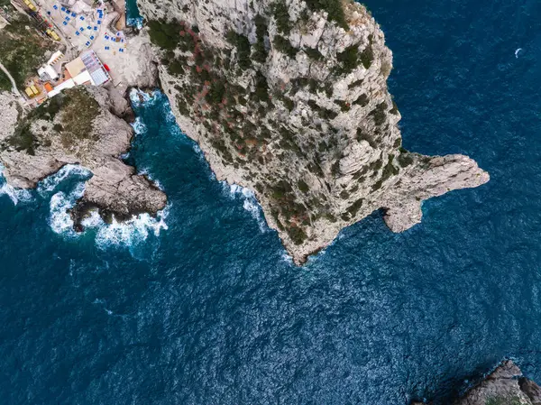 İtalya 'nın Capri Adası' ndaki kayalık bir uçurum perspektifi derin mavi Akdeniz sularıyla çevrilidir. Nadir bulunan bitki örtüsü ve plaj bölgesi görülebilir..