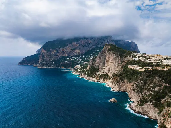Capri Adası 'nın kıyı şeridinde Tyrhenian Denizi' nden yükselen kayalıklar, beyazlatılmış binalar, yemyeşil alanlar ve dağ tepelerinin üzerindeki bulutlar yer alır..