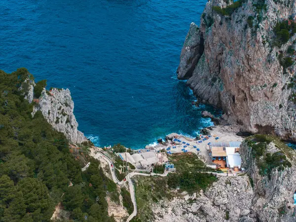 İtalya, Capri Adası 'ndaki sahil manzarası, sarp kayalıklar, canlı mavi sular, şemsiyeli küçük bir sahil ve dolambaçlı yollar boyunca yemyeşil..
