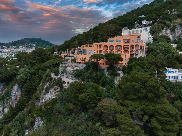 Kemerli pencere ve terasları olan turuncu bir villa Capri, İtalya 'da yeşil bir yamaçta, taş bir köprünün yanında ve pembe mavi gökyüzünün altındaki engebeli kayalıklarda bulunur..