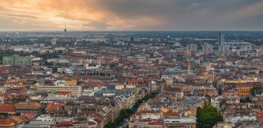 Macaristan, Budapeşte 'nin havadan görünüşü, kırmızı kiremitli çatılar, süslü cepheler ve dramatik bir günbatımı gökyüzü altında uzun bir telekomünikasyon kulesi..