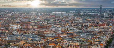 Macaristan 'ın Budapeşte kentinin havadan görünüşü, kırmızı ve turuncu çatılar, modern bir stadyum, yüksek bir gökdelen ve bulutların arasından yükselen güneş ışığı..