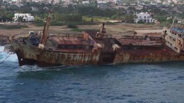 Kıbrıs 'ın kayalık kıyıları yakınlarındaki Edro III enkazının hava temizliği, paslı gövde, mavi sular, engebeli uçurumlar, kumlu kıyılar ve yakınlardaki binaları gösteriyor.