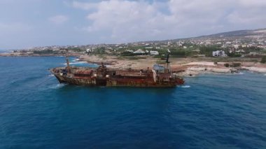 Kıbrıs 'ın kayalık kıyıları yakınlarındaki Edro III enkazının hava manzarası, dalgalar, kayalıklar, kumlu alanlar, bitki örtüsü ve bulutlu bir gökyüzünün altındaki uzak bir kasaba..
