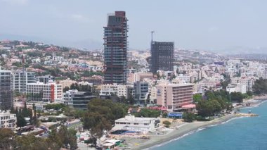 Kıbrıs Rum Kesimi 'nin Limasol kentinin havadan görünüşü, kumlu bir sahil, dalga kırıcıları olan berrak sular, modern yüksek binalar ve arka planda kayan tepeler gösteriyor..
