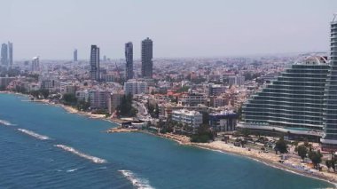 Kıbrıs Rum Kesimi 'nin Limasol kentinin hava görüntüsü, modern binalar, kırmızı çatılı evler, vinçler, kıyı şeridi, yeşillik ve tepecikli araziyi yavaş bir tavada gösteriyor..