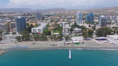 Kıbrıs 'ın Limasol kentinin havadan görünüşü, yüksek binalar, beyaz binalar, sıralanmış bir ağaç gezinti güvertesi, turkuaz sular, bir plaj ve yuvarlanan tepeleri gösteriyor..