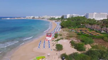 Kıbrıs 'taki Ayia Napa plajının yavaş bir kısmı, altın kum, turkuaz sular, güneşlik yataklar, cankurtaran kulesi ve yeşillikli tatil köyü binalarını gösteriyor..