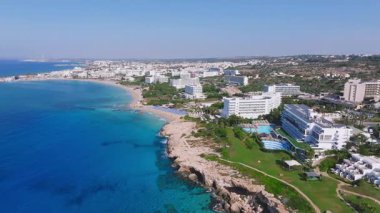 Kıbrıs Rum Kesimi 'nin Ayia Napa kentinin hava manzarası, tatil beldeleri, mavi havuzlar, kayalık uçurumlar, turkuaz sular ve pürüzsüz dönüm hareketiyle yemyeşil alanlar gösteriyor.