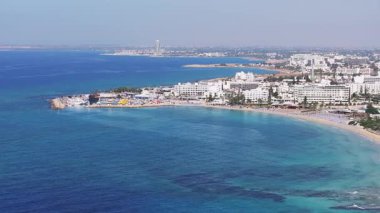 Kıbrıs Rum Kesimi 'nin Ayia Napa kentinin hava manzarasında turkuaz sular, güneşli kumlu plajlar, modern tatil beldeleri, yemyeşil alanlar ve kentsel yerleşim alanları görülüyor..