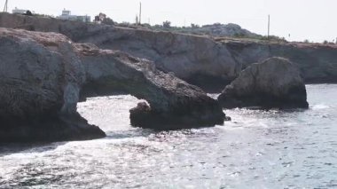 Kıbrıs Rum Kesimi 'nin Ayia Napa kentindeki engebeli uçurumlar ve berrak mavi sular. Yumuşak dalgalar kıyıya vurur palmiye ağaçları ve arka planda beyaz binalar.