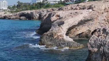 Kıbrıs Rum Kesimi 'nin Ayia Napa kentinin hava manzarası kayalık kıyı şeridi, turkuaz sular, güneşli kumsallar ve beyaz tatil köyleri binaları arasında dönüyor..