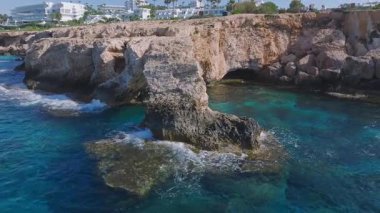 Kıbrıs Rum Kesimi 'nin Ayia Napa kentindeki Aşk Kemeri' nin turkuvaz suları ve engebeli uçurumlara çarpan dalgalarla hava görüntüsü. Kamera çeşitli açıları göstermek için hareket eder.