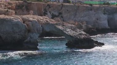Ayia Napa 'nın kireçtaşı kayalıkları, doğal kemerler, turkuaz suları olan mağaralar ve arka planda bir tatil köyü. Dalgalar yavaşça aşağı hareket eder.