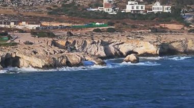 Ayia Napa 'nın Aşk Kemeri kaya oluşumu, engebeli uçurumlar ve Akdeniz dalgaları. Modern beyaz villalar arka planda beliriyor, doğayla iç içe..