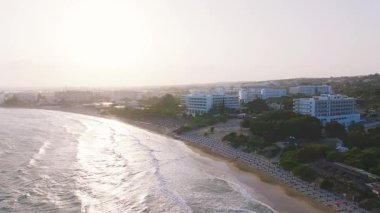 Gün batımında Ayia Napa 'nın hava görüntüsü, güneşli yataklar, şemsiyeler, temiz sular, kumlu kıyılar, tatil beldeleri ve kentsel manzaralı sahili gösteriyor..