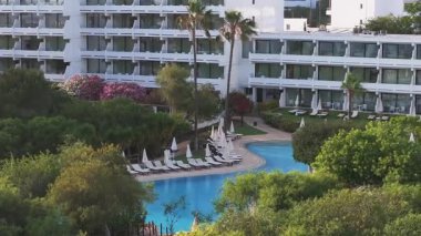 Kıbrıs Rum Kesimi 'nin Ayia Napa kentindeki bir tatil köyünün hava manzarasında beyaz bir bina, büyük bir havuz, şezlonglar, şemsiyeler ve canlı çiçekli çalılar görülüyor..