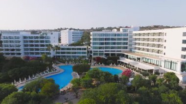 Kıbrıs Rum Kesimi 'nin Ayia Napa kentindeki havuz manzarası, geniş bir havuz, modern beyaz binalar, yemyeşil yemyeşil ve yumuşak güneş ışığında kademeli dönüm hareketi.