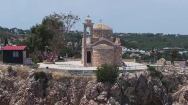 Kıbrıs Rum Kesimi 'nin Ayia Napa kentindeki Aziz Elias Kilisesi, patika üzerinde bayraklar bulunan kayalık bir tepenin üzerinde bulunuyor. Ziyaretçiler bölgeyi çevreleyen yeşil alanları ve binaları keşfediyorlar.