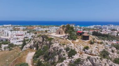 Kıbrıs Rum Kesimi 'nin Ayia Napa kentinin hava manzarasında bir tepe üzerinde Aziz Elias Kilisesi, beyazlatılmış binalar, kayalık arazi ve mavi Akdeniz yer alıyor..