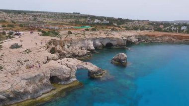 Love Arch kaya oluşumu, turkuaz sular, kayalık arazi, dağınık binalar ve Kıbrıs Rum Kesimi yakınlarındaki yemyeşil arazi üzerinde yumuşak bir insansız hava aracı uçuşu.