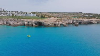 Kıbrıs Rum Kesimi 'nin Ayia Napa kentinin hava görüntüsü, kireçtaşı kayalıkları, berrak mavi sular, yeşil çimenli beyaz bir tatil köyü ve araziyi keşfeden insanlar gösteriyor..