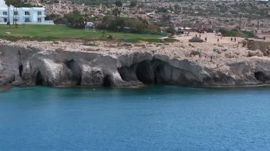 Kıbrıs 'ın Ayia Napa kentindeki sakin kıyı sahnesinde Aşk Kemeri, kayalık kayalıklar, mağaralar, berrak sular ve hareket halindeki yeşil şişme bir su aracı yer alıyor..