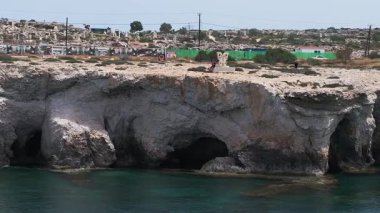 Kıbrıs Rum Kesimi 'nin Ayia Napa kentindeki engebeli uçurumlar ve berrak turkuaz sular, kenarda yürüyen insanlar ve arka planda küçük bir yerleşim yeri.