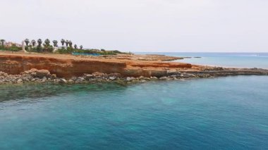 Ayia Napa 'nın kıyı şeridini kırmızımsı kahverengi uçurumlar, turkuaz sular, palmiye ağaçları ve arka planda dağınık tatil beldeleri ile gösteren hava manzarası.