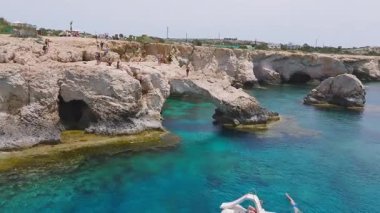 Kıbrıs 'ın Ayia Napa kentinde doğal kaya oluşumu Love Arch. Ziyaretçiler kayalıklarda toplanıyor, bir yüzücü aşağıdaki berrak turkuaz denize dalıyor..