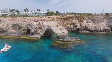 Kıbrıs Rum Kesimi 'nin Ayia Napa kentindeki turkuaz sular, engebeli uçurumlar, deniz mağaraları ve kemerler. Ziyaretçiler yukarıdaki kayalık araziyi gezerken bir tekne yavaşça hareket eder..