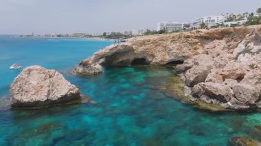 Kıbrıs Rum Kesimi 'nin Ayia Napa kentindeki Aşk Kemeri' nin hava görüntüsü, kemer, turkuaz sular, tatil beldesi binaları ve arka planda yemyeşil alanlar üzerinde yürüyen insanlar..