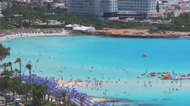 Kıbrıs 'taki Ayia Napa Nissi plajının hava manzarasında turkuaz sular, yüzücüler, kanocular, bir tekne, modern tatil beldeleri ve yemyeşil alanlar görülüyor..