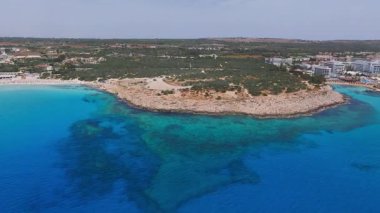 Kıbrıs Rum Kesimi 'nin Ayia Napa kentinin hava manzarasında turkuaz sular, kayalık kıyılar, plaj şemsiyeleri, park edilmiş araçlar ve kumlu arazi ortasında tatil beldeleri görülüyor..