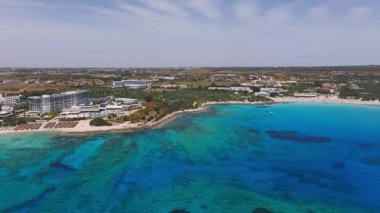 Kıbrıs Rum Kesimi 'nin Ayia Napa kentinin havadan görünüşü, hilal şeklinde bir plaj, turkuaz sular, yemyeşil alanlar ve pürüzsüz insansız hava aracı hareketine sahip tatil merkezleri gösteriyor..