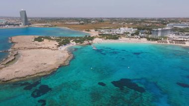 Kıbrıs Rum Kesimi 'nin Ayia Napa kentinin pürüzsüz havası turkuaz sular, kumlu plajlar, tatil beldeleri, bir lagün ve kıyıya yakın tekneleri olan bir rıhtım gösteriyor..
