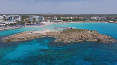 Kıbrıs Rum Kesimi 'nin Ayia Napa kentinin hava manzarası, turkuvaz suları, mavi şemsiyeler, kayalık çıkıntılar ve yakın tatil beldeleri ile Nissi Plajı' nı geniş çaplı bir hareketle gösteriyor..