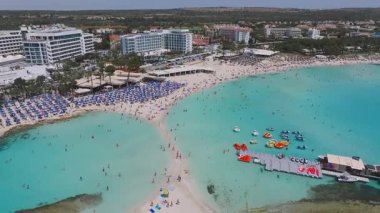 Kıbrıs Rum Kesimi 'nin Ayia Napa Nissi Plajı' nın hava manzarasında turkuaz sular, kayalık bir yarımada, sahil tatil beldeleri ve hareketli kıyı şeridi yakınlarındaki tekneler görülüyor..