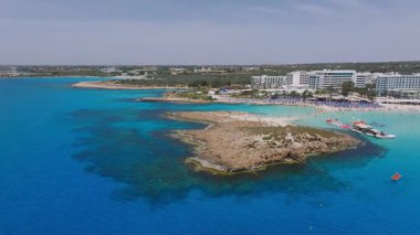 Kıbrıs Rum Kesimi 'nin Ayia Napa kentinin havadan görünüşü turkuaz sular, kırmızı ve sarı tekneler, kumlu Nissi plajları, beyaz tatil beldeleri ve hareketli yemyeşil alanları gösteriyor..