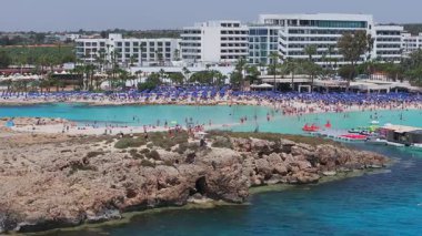 Kıbrıs Rum Kesimi 'nin Ayia Napa kentinin hava manzarasında turkuaz sular, kayalık kıyılar, beyaz bir şapel, su parkı, canlı plajlar ve kentsel çevre görülüyor..