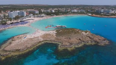 Kıbrıs Rum Kesimi 'nin Ayia Napa kentinin havadan görünüşü turkuaz suları, kumlu Nissi plajları, sahil tatil beldeleri, güneşlik yatakları ve denizde hareket eden tekneleri gösteriyor..