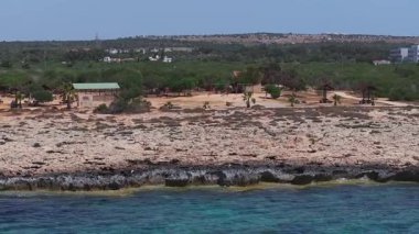 Kıbrıs Rum Kesimi 'nin Ayia Napa kentinin hava manzarası, berrak turkuaz suları, modern beyaz tatil beldeleri ve kuru arazide seyrek bitki örtüsü bulunan kayalık bir kıyı şeridini gösteriyor..