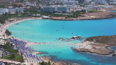 Kıbrıs Rum Kesimi 'nin Ayia Napa kentinin havadan görünüşü turkuaz suları, kayalık bir adalı kumlu Nissi plajı, rıhtıma yanaşmış bir tekne ve hareket halindeki bir sürat teknesi gösteriyor..