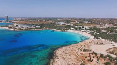 Kıbrıs Rum Kesimi 'nin Ayia Napa kentinin hava manzarasında turkuaz sular, kumlu plajlar, kayalık arazi, yemyeşil ve arka planda bir grup tatil köyü görülüyor..