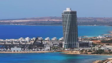 Kıbrıs 'taki Ayia Napa marinasının hava manzarası, sergilenen yatlar, kıvrımlı bir dalgakıran, modern liman binaları ve kristal berrak mavi sular.