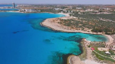 Kıbrıs Rum Kesimi 'nin Ayia Napa kentinin hava manzarası turkuaz suları, kayalık kıyıları, Aşk Kemeri, tatil beldeleri, Nissi Sahili, Landa Sahili ve hafif dalga hareketlerini gösteriyor..