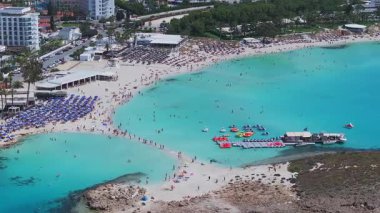 Kıbrıs Rum Kesimi 'nin Ayia Napa kentindeki Nissi Sahili' nin hava manzarasında turkuaz sular, güneşlenenler, tekneler, jet ski 'ler, kayalıklar ve yakınlardaki tatil beldeleri görülüyor..