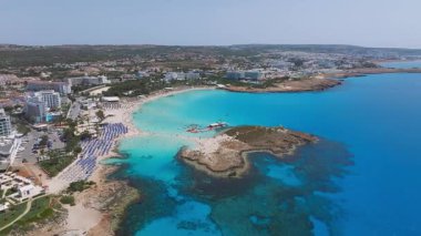Kıbrıs 'ın Ayia Napa kentinin hava manzarası, Nissi Sahili, turkuaz sular, modern tatil beldeleri, kayalık çıkıntılar, tekneler ve canlı sahil faaliyetlerinin sergilendiği.