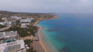 Kıbrıs 'ın Ayia Napa kentinin hava manzarası, turkuaz sular, kumlu plajlar, tatil beldeleri, kayalık çıkıntılar ve kıyı boyunca renkli şemsiyeler sergileniyor.