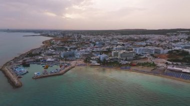 Günbatımında Kıbrıs 'ın Ayia Napa kentinin hava manzarası. Beyaz binalar, tatil beldeleri, havuzlar ve sakin sulara sahip sahil şeridi ağır çekimde yakalanır..